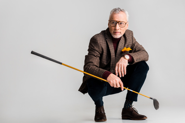 trendy mature man in glasses holding golf club and while sitting on grey - Foto, Bild
