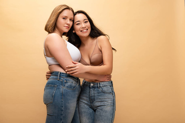 pensive overweight young woman and smiling slim asian girl hugging while looking at camera isolated on beige, body positivity concept - Photo, Image