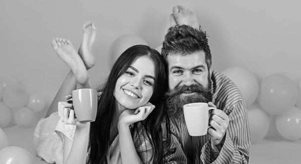 Man and woman on smiling faces lay, pink background. Couple in love drink coffee in bed. Couple relax in morning with coffee. Family tradition concept. Man and woman in domestic clothes, pajamas - Zdjęcie, obraz