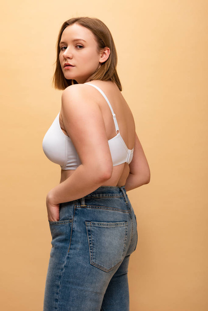 pretty overweight girl in blue jeans and bra looking at camera isolated on beige, body positivity concept - Foto, Bild