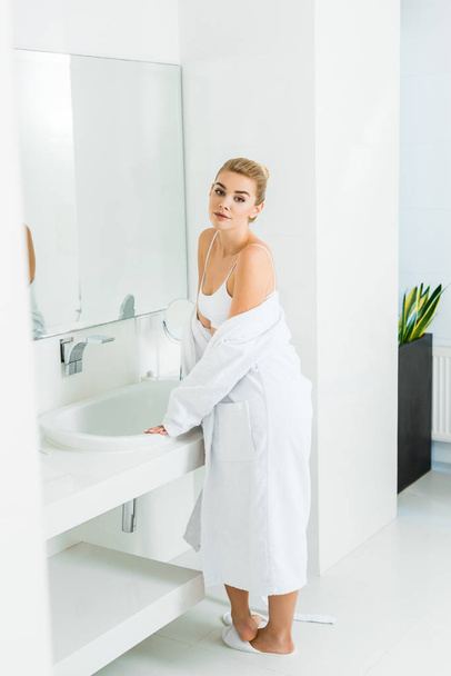 hermosa y rubia mujer en sujetador y albornoz blanco mirando a la cámara en el baño
  - Foto, Imagen