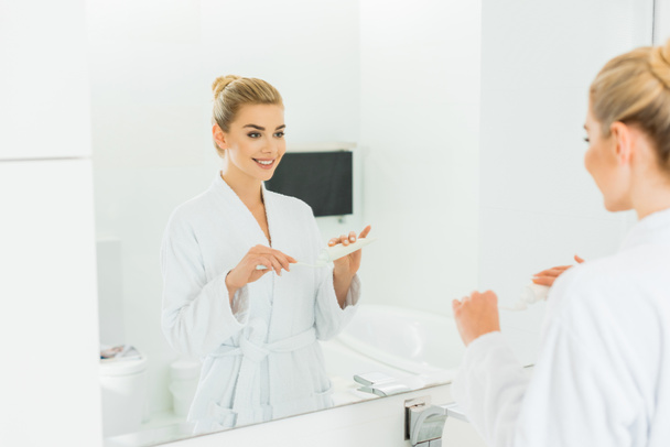 enfoque selectivo de la mujer sonriente en albornoz aplicando pasta de dientes en el cepillo de dientes en el baño
  - Foto, Imagen