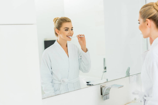 selective focus of woman in white bathrobe brushing teeth and looking at mirror  - Fotó, kép