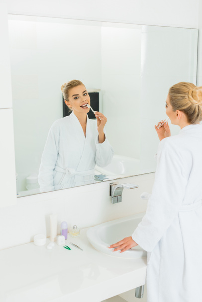 bela mulher em roupão de banho branco escovando os dentes e olhando para o espelho
  - Foto, Imagem