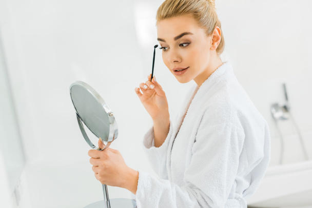 attraktive Frau im weißen Bademantel, die Augenbrauen putzt und in den Spiegel schaut  - Foto, Bild