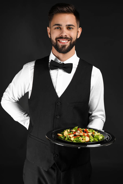 Knappe kelner met frisse salade op donkere achtergrond - Foto, afbeelding