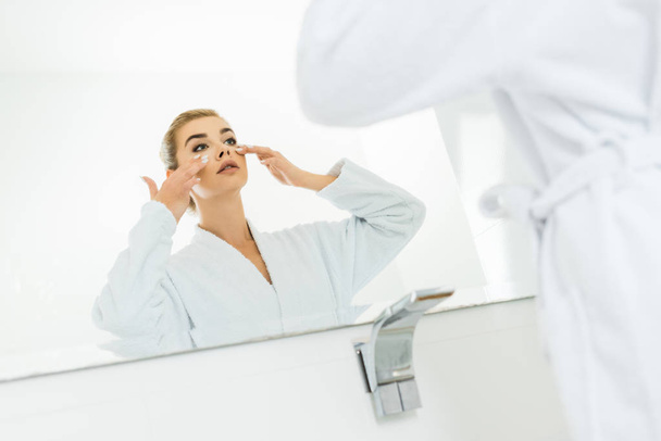 selectieve aandacht van vrouw in witte badjas gezichtscrème in badkamer toe te passen - Foto, afbeelding