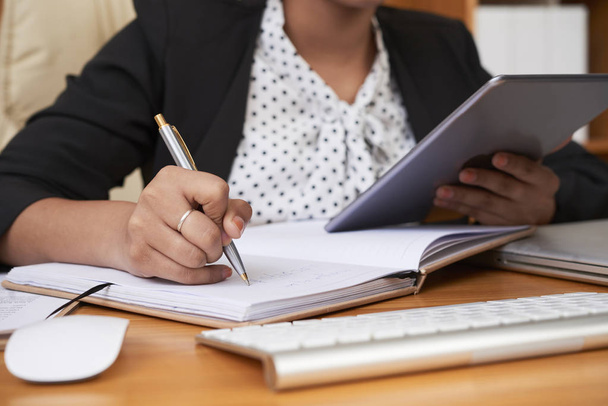 Close-up de trabalhadora de escritório escrevendo novas ideias de negócios em seu bloco de notas enquanto usa tablet digital em seu local de trabalho no escritório
 - Foto, Imagem
