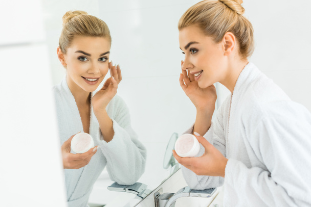 selective focus of attractive and blonde woman in white bathrobe applying face cream and looking at mirror  - Photo, Image