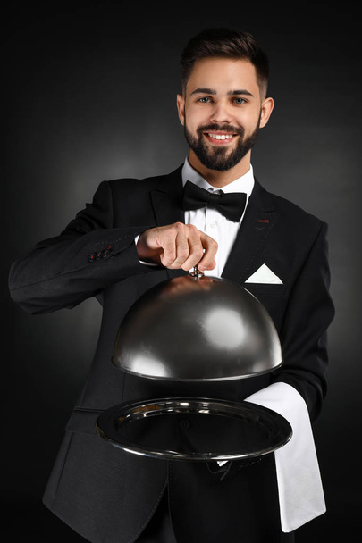 Knappe kelner met lade en cloche op donkere achtergrond - Foto, afbeelding