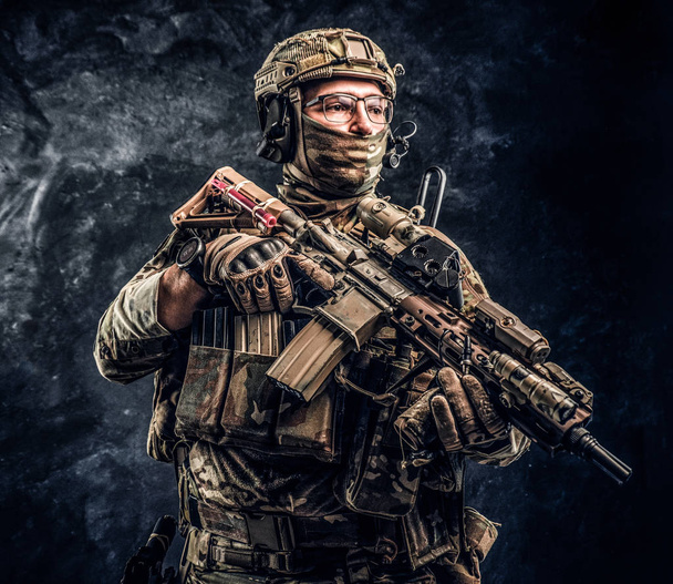 Повністю обладнаний солдат у камуфляжній формі, що тримає штурмову гвинтівку. Студійне фото на тлі темної стіни
 - Фото, зображення