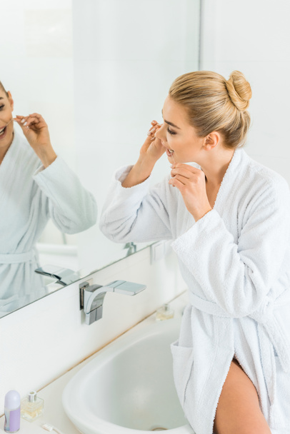 Selektivní fokus atraktivní a blond žena v bílém županu čištění zubů s dentální nit - Fotografie, Obrázek