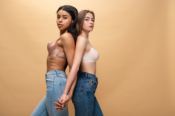 two beautiful multicultural women standing back to back and holding hands isolated on beige  - Фото, изображение