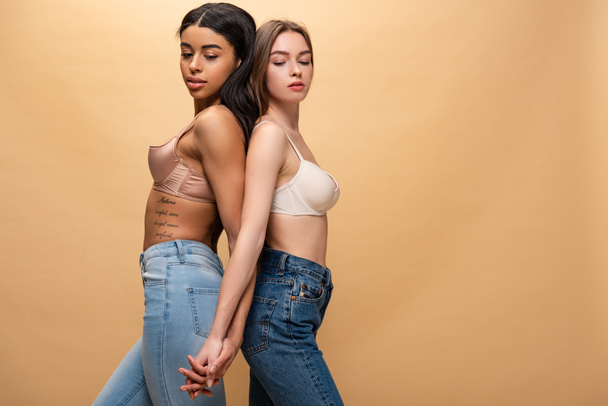 two pensive multicultural girls holding hands while standing back to back isolated on beige  - Foto, Bild