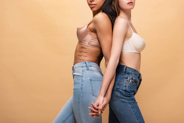 abgeschnittene Ansicht zweier multikultureller Mädchen, die sich Händchen halten, während sie Rücken an Rücken isoliert auf beige stehen  - Foto, Bild