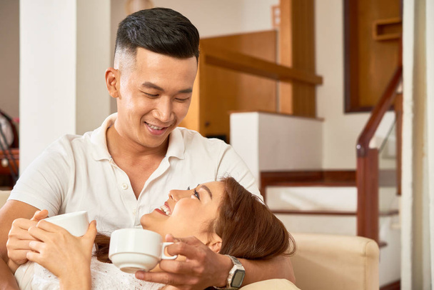 Glimlachend Aziatische paar omarmen en samen koffie drinken terwijl u ontspant op sofa thuis - Foto, afbeelding