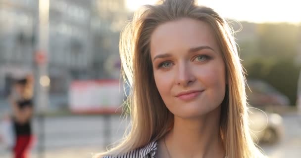 A young lady is smiling at the camera on the street. The sun is shining and sun rays blink the camera. Close-up portrait. - Кадры, видео