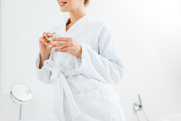 cropped view of woman in white bathrobe holding perfume in bathroom  - Фото, зображення