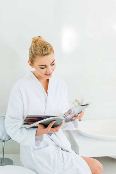 attractive and smiling woman in white bathrobe reading magazine in bathroom  - Фото, зображення