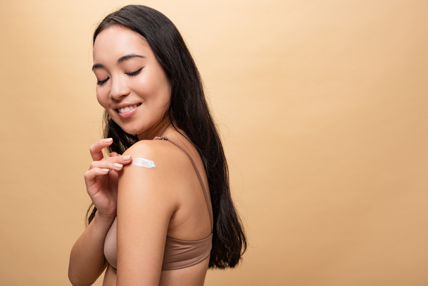 feliz mulher asiática aplicando creme cosmético no ombro isolado no bege
  - Foto, Imagem
