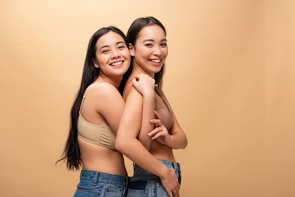 two smiling multicultural young women hugging while posing at camera isolated on beige, body positivity concept - 写真・画像