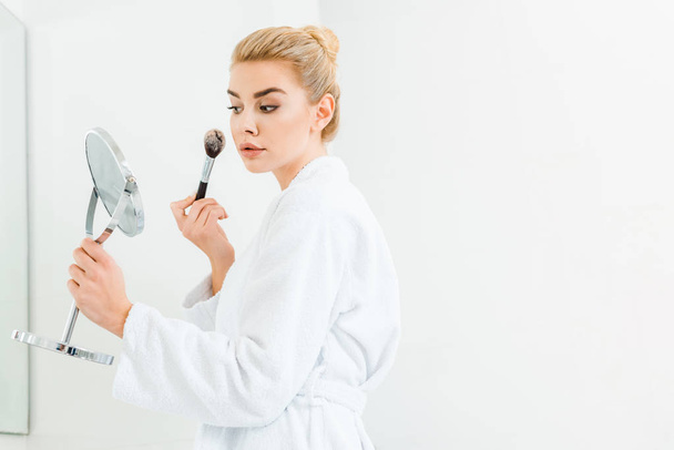 beautiful and blonde woman in white bathrobe using cosmetic brush in bathroom  - Фото, зображення