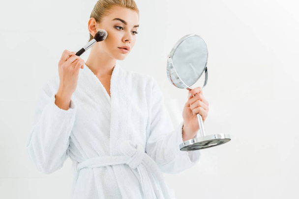 attractive and blonde woman in white bathrobe using cosmetic brush and holding mirror  - Foto, immagini