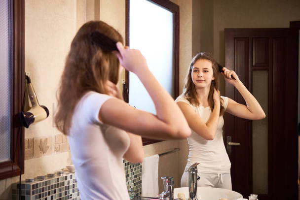 Fiatal lány nézett tükörbe, és ecset hosszú haja a fürdőszobában hajkefe - Fotó, kép