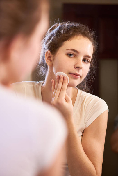 Jeune femme regardant miroir et nettoyant son visage du maquillage avec du coton tout en restant debout dans la salle de bain
 - Photo, image