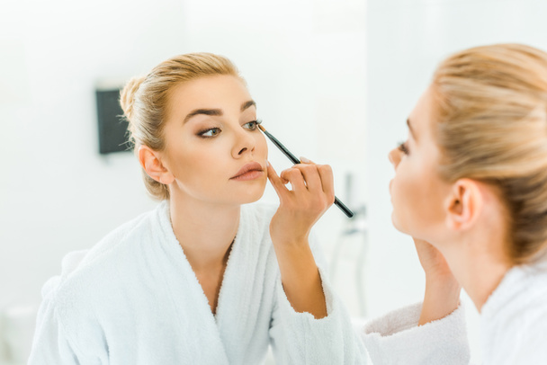 selective focus of woman in white bathrobe applying eyeshadow with cosmetic brush - Photo, Image