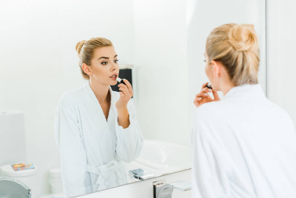 selective focus of woman in white bathrobe applying lipstick and looking at mirror in bathroom  - 写真・画像