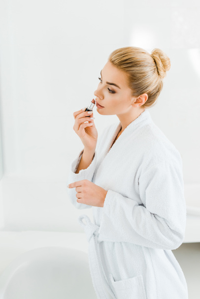 beautiful and blonde woman in white bathrobe applying lipstick in bathroom  - Photo, Image