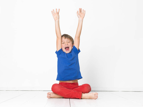 six year old boy in red trousers and blue t-shirt shouting sitting with crossed legs and raised arms on white floor - Foto, imagen
