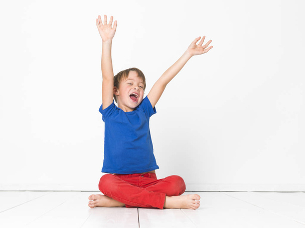 six year old boy in red trousers and blue t-shirt shouting sitting with crossed legs and raised arms on white floor - Photo, Image