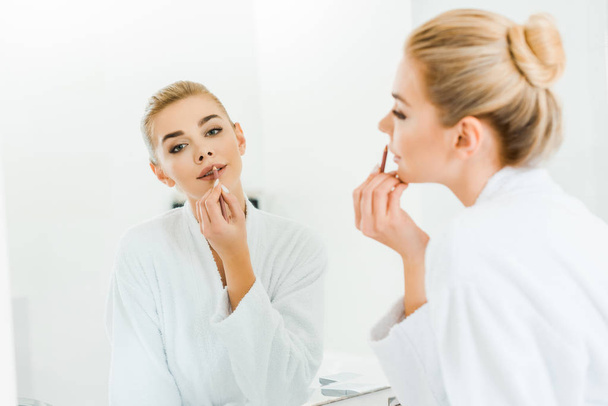 selective focus of beautiful and blonde woman in white bathrobe applying lip liner and looking at mirror  - Fotó, kép