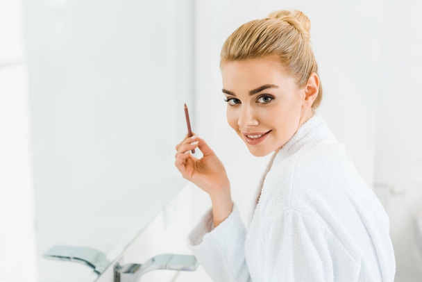 beautiful and smiling woman in white bathrobe holding lip liner and looking at camera  - Foto, immagini