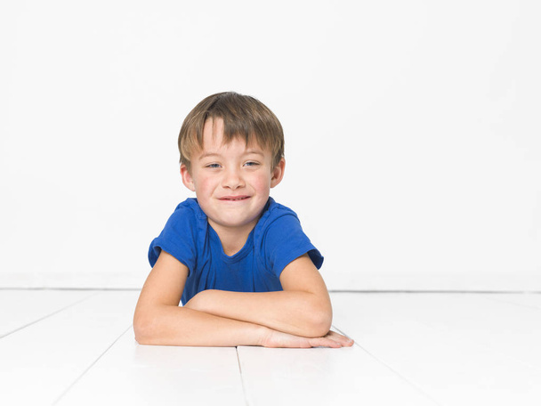 bambino di sei anni in t-shirt blu guardando la macchina fotografica e sdraiato con le braccia incrociate sul pavimento bianco
 - Foto, immagini