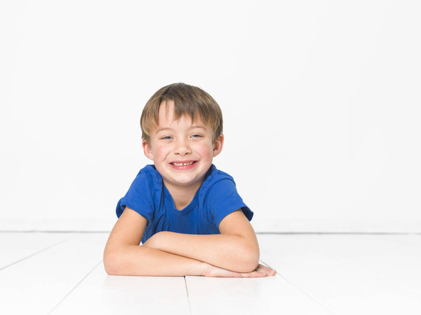 six year old boy in blue t-shirt looking at camera and lying with crossed arms on white floor - Фото, зображення