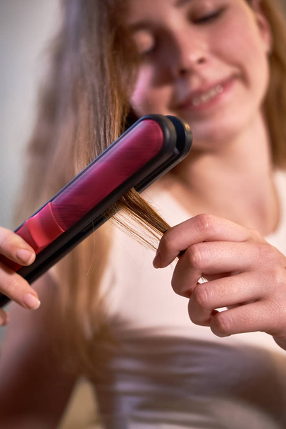 Close-up of young woman holding hair straightener and straightening her long hair herself - Fotoğraf, Görsel