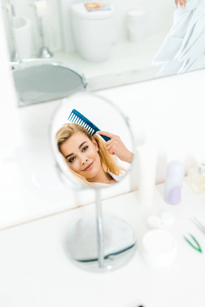 selective focus of beautiful and smiling woman holding comb and looking at mirror  - Foto, Imagem