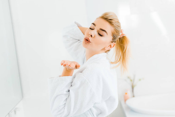 beautiful woman in white bathrobe playing with hair and blowing air kiss in bathroom  - Foto, Imagem