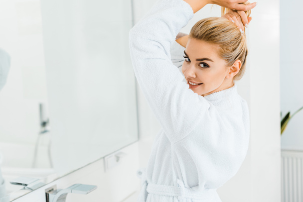 beautiful woman in white bathrobe playing with hair and looking at camera in bathroom  - Photo, Image