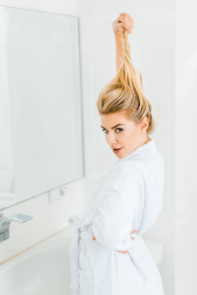 attractive woman in white bathrobe playing with hair and looking at camera in bathroom  - Foto, immagini
