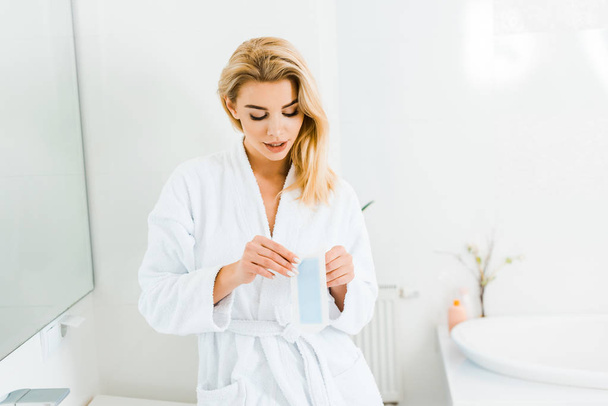 aantrekkelijke vrouw in witte badjas holding ontharing stripe in badkamer  - Foto, afbeelding