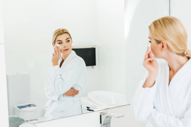 selective focus of beautiful and blonde woman in white bathrobe using cotton pad and looking at mirror  - Фото, изображение