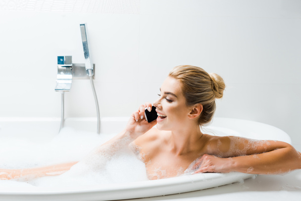 attractive and smiling woman taking bath with foam and talking on smartphone in bathroom  - Fotografie, Obrázek