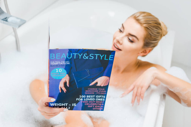 selective focus of attractive and blonde woman taking bath with foam and reading beauty and style magazine in bathroom  - Foto, Bild