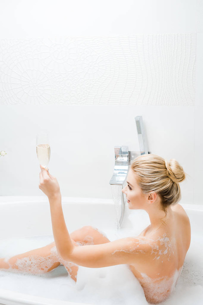 beautiful and smiling woman taking bath and holding champagne glass in bathroom  - Фото, зображення