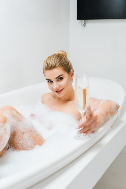 foco seletivo da mulher bonita e sorridente tomando banho e segurando copo de champanhe no banheiro
  - Foto, Imagem