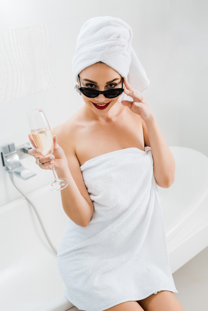 beautiful and smiling woman in sunglasses and towels holding champagne glass and looking at camera in bathroom  - Foto, Imagem